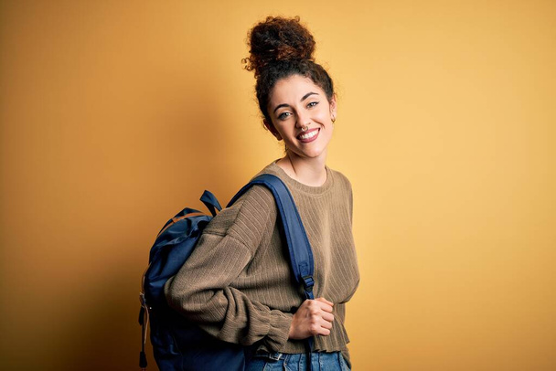 Mladá krásná studentka s kudrnatými vlasy a piercing na sobě batoh s veselou tváří stojící a usmívající se s sebevědomým úsměvem ukazující zuby - Fotografie, Obrázek