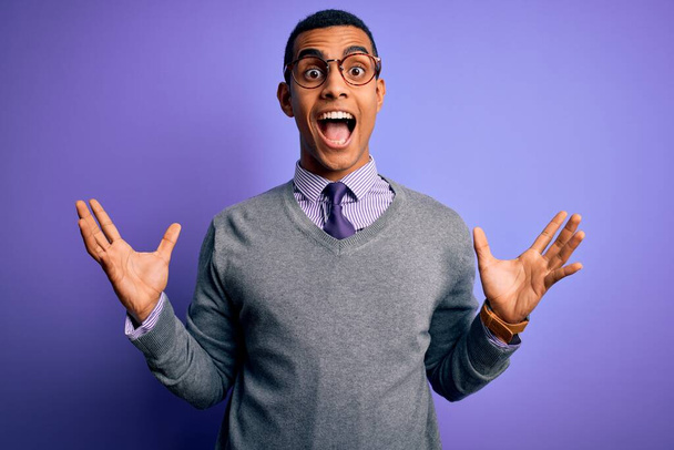 Hombre de negocios afroamericano guapo con gafas y corbata sobre fondo púrpura celebrando loco y sorprendido por el éxito con los brazos levantados y los ojos abiertos gritando emocionado. Concepto ganador
 - Foto, imagen