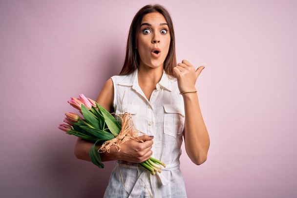Jeune belle femme brune tenant bouquet de fleurs de tulipes sur fond rose Surprise pointant du doigt sur le côté, bouche ouverte expression étonnée
. - Photo, image
