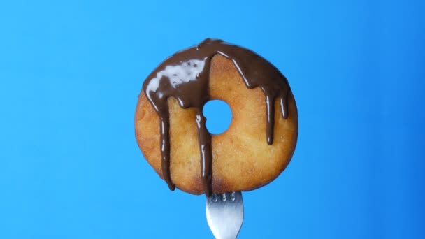 Big round donut on a fork on which chocolate icing glaze flows on a blue background - Footage, Video