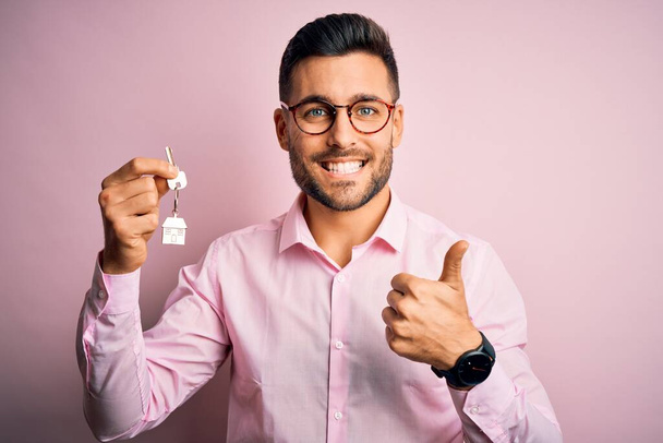 Young real estate business man holding new house keys over pink background happy with big smile doing ok sign, thumb up with fingers, excellent sign - Foto, Imagem