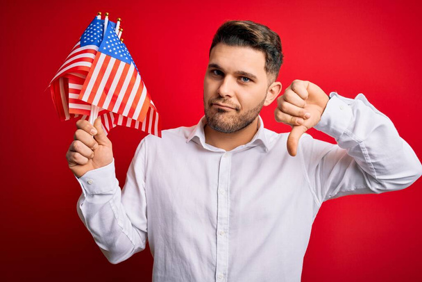 Nuori sinisilmäinen mies, jolla on Amerikan yhdysvaltojen lippu punaisella, eristetyllä taustalla vihaiset kasvot, kielteinen merkki, joka osoittaa vastenmielisyyttä peukalot alhaalla, hylkäyskäsite
 - Valokuva, kuva
