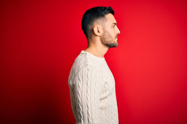 Young handsome man wearing casual white sweater standing over isolated red background looking to side, relax profile pose with natural face with confident smile. - Φωτογραφία, εικόνα
