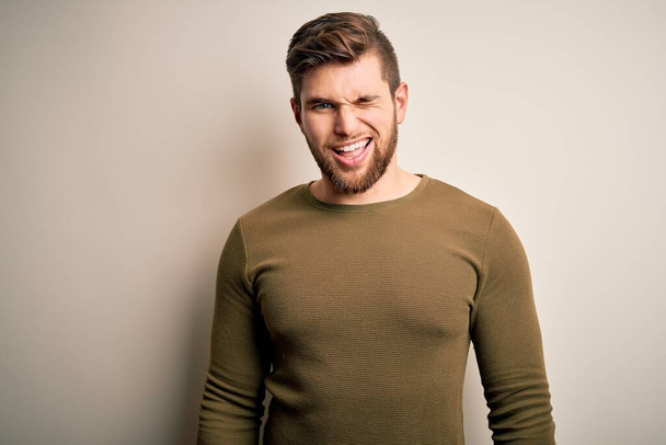 Young blond man with beard and blue eyes wearing green sweater over white background winking looking at the camera with sexy expression, cheerful and happy face. - Photo, Image