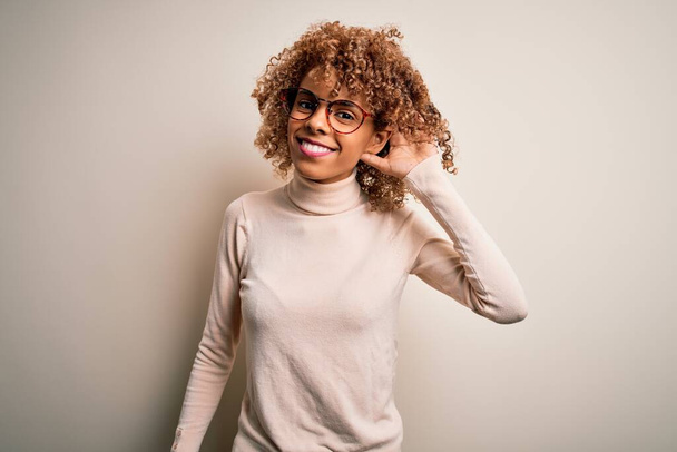 Młoda Afroamerykanka w swetrze i okularach na białym tle, uśmiechnięta z ręki na ucho, słuchająca plotek i plotek. Koncepcja głuchoty. - Zdjęcie, obraz