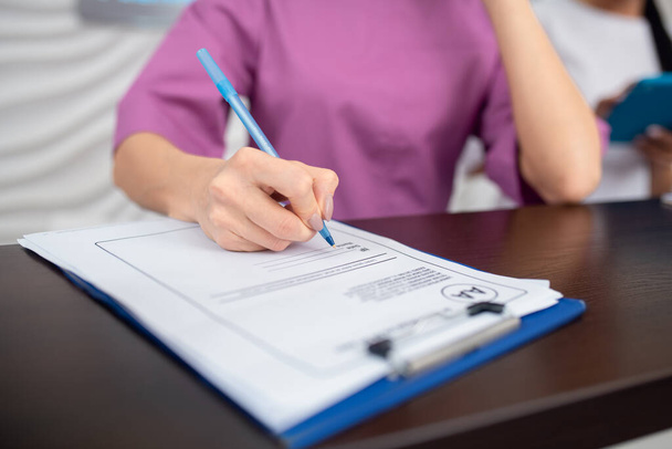 Receptionist φορώντας μωβ στολή κρατώντας στυλό, ενώ κρατώντας σημειώσεις - Φωτογραφία, εικόνα