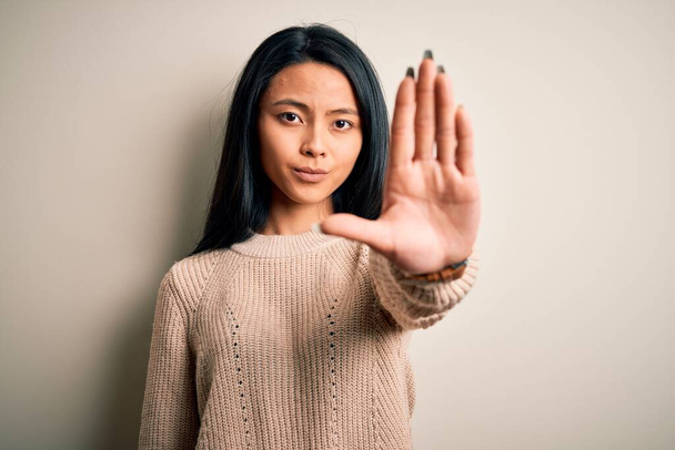 孤立した白い背景の上にカジュアルなセーターを着て若い美しい中国の女性は手の手のひらで歌うのを停止します。顔に負と深刻なジェスチャーで警告式. - 写真・画像