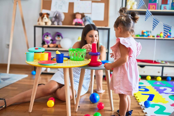 Young beautiful teacher and toddler building pyramid using hoops on the table at kindergarten - Photo, image