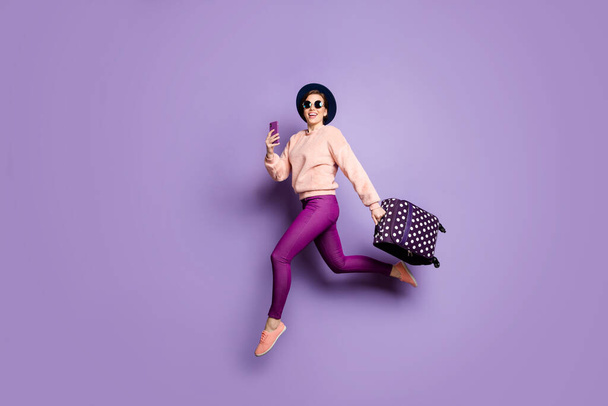 Full body profile photo of funny lady jumping up rushing airport hold telephone carry luggage tourist wear cap sun glasses trendy pink pullover trousers isolated purple color background - Foto, imagen