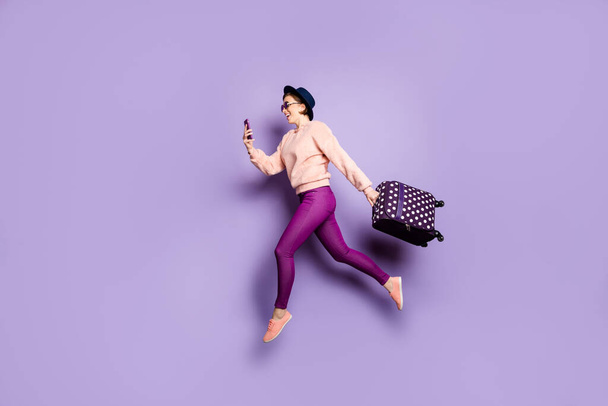 Teljes hossz profil fotó vicces hölgy jumping magas séta repülőtér néz telefon hordozza poggyász viselet sapka napszemüveg alkalmi trendi rózsaszín pulóver nadrág elszigetelt lila háttér - Fotó, kép