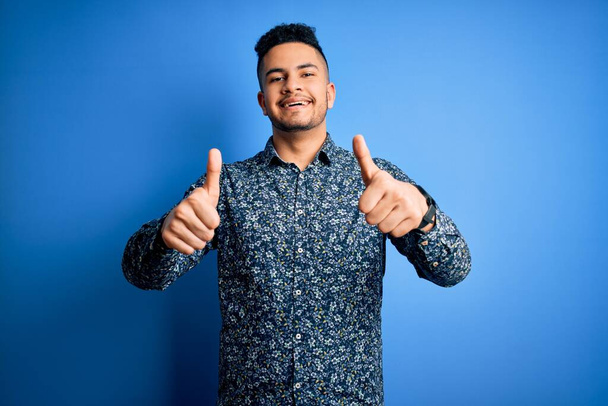 Young handsome man wearing casual shirt standing over isolated blue background success sign doing positive gesture with hand, thumbs up smiling and happy. Cheerful expression and winner gesture. - Photo, Image