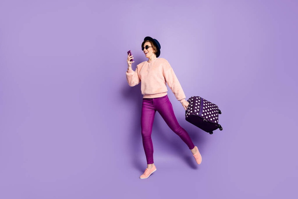 Full size profile photo of nice lady walking registration hold telephone carry bag waiting hall wear hat sun glasses casual trendy pink sweater trousers isolated purple color background - Foto, imagen
