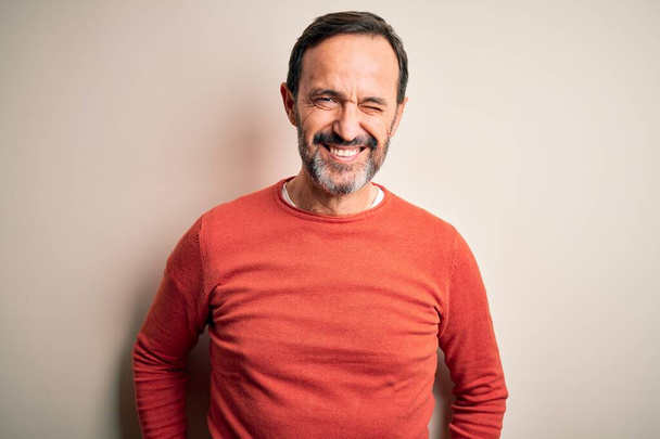 Middle age hoary man wearing casual orange sweater standing over isolated white background winking looking at the camera with sexy expression, cheerful and happy face. - Photo, Image
