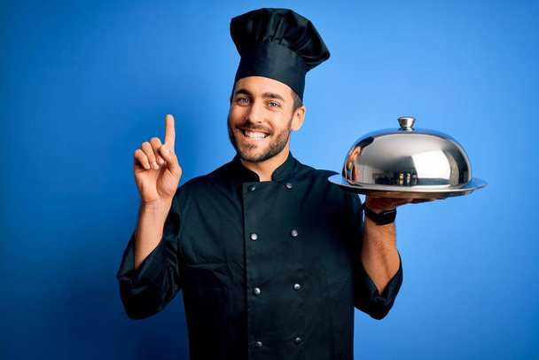 Młody kucharz z brodą w mundurze trzymający tacę z kopułą na niebieskim tle zaskoczony pomysłem lub pytaniem wskazującym palcem na szczęśliwą twarz, numer jeden - Zdjęcie, obraz