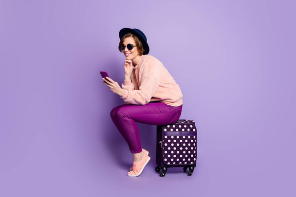 Full length profile photo of pretty lady waiting registration flight hold telephone sitting on bag wear hat sun specs casual trendy pink sweater pants isolated purple color background - Fotoğraf, Görsel