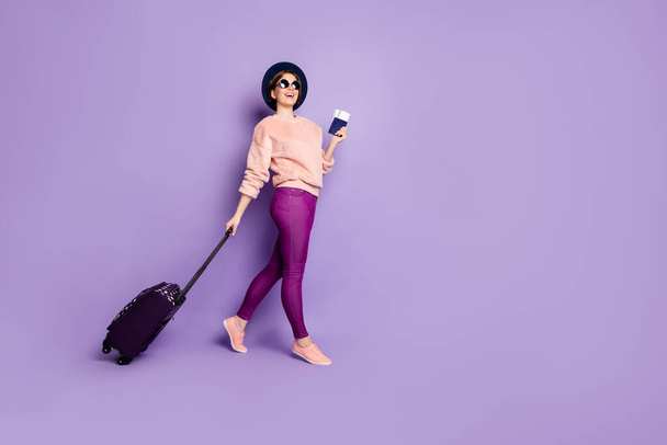 Full size photo of funny lady vacation walking registration flight hold ticket passport rolling bag wear hat sun specs casual trendy pink sweater pants isolated purple color background - Foto, Bild