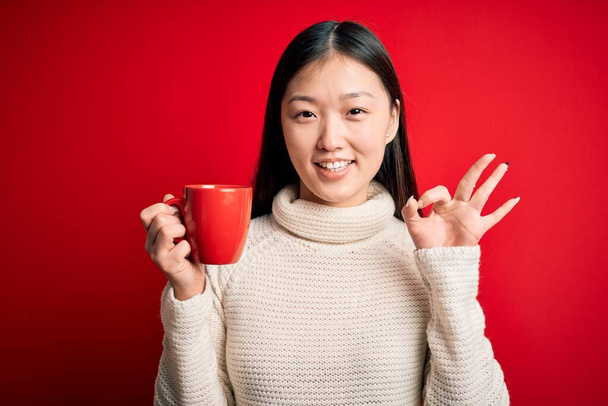Young asian woman drinking a cup of hot coffee over isolated red background doing ok sign with fingers, excellent symbol - Fotó, kép