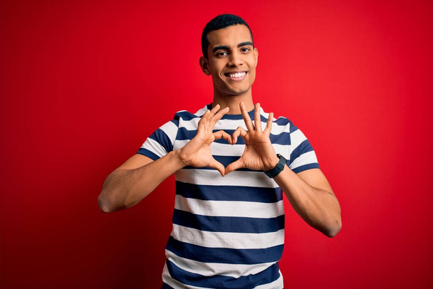 Hombre afroamericano guapo vistiendo camiseta casual a rayas de pie sobre fondo rojo sonriendo en el amor haciendo forma de símbolo del corazón con las manos. Concepto romántico
. - Foto, imagen