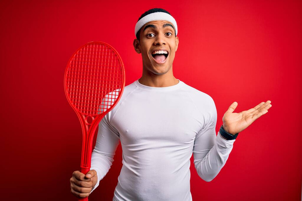 Genç ve yakışıklı Afro-Amerikan sporcusu kırmızı arka planda raket kullanarak tenis oynuyor. Çok mutlu ve heyecanlı, kazanan yüz ifadesi zaferi büyük bir gülümsemeyle kutluyor ve ellerini kaldırıyor. - Fotoğraf, Görsel