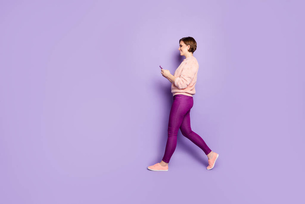 Full length profile photo of pretty lady holding telephone chatting with friends walking down street wear casual stylish pink pullover trousers isolated purple color background - Fotó, kép