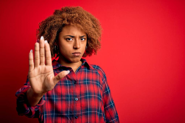 Jonge mooie Afro-Amerikaanse afro vrouw met krullend haar dragen casual shirt doen stoppen met zingen met palm van de hand. Waarschuwingsuitdrukking met negatief en ernstig gebaar op het gezicht. - Foto, afbeelding
