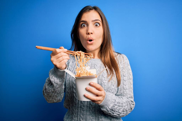 Young woman eating asian noodles from take away box using chopstick over blue background scared in shock with a surprise face, afraid and excited with fear expression - Фото, зображення