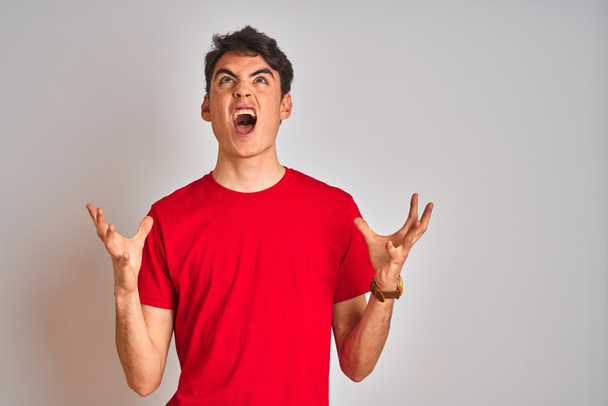 Tiener jongen draagt rood t-shirt over witte geïsoleerde achtergrond gek en gek schreeuwen en schreeuwen met agressieve uitdrukking en armen omhoog. Frustratieconcept. - Foto, afbeelding