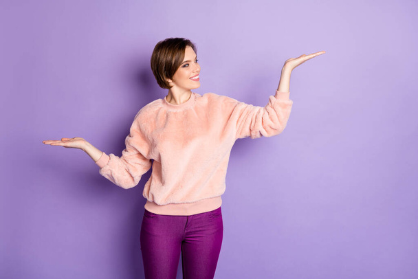 Portrait of cheerful positive girl hold hand raise palm up compare offer try choose promotion adverts wear pastel pullover isolated over violet color background - Foto, afbeelding