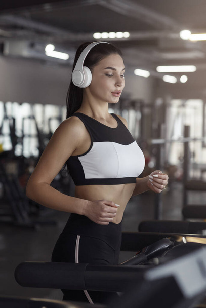 Pretty girl with headphones jogging on treadmill in gym - Fotó, kép