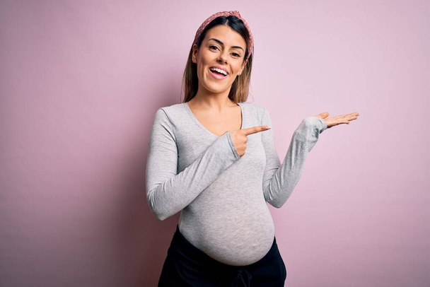 Mladá krásná brunetka žena těhotná čeká dítě přes izolované růžové pozadí ohromen a usmívá se na kameru při prezentaci s rukou a ukazuje prstem. - Fotografie, Obrázek