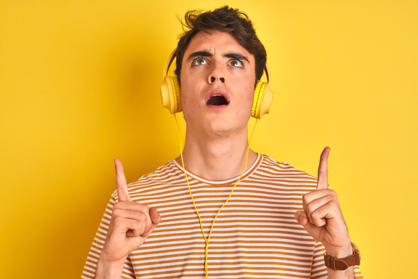 Teenager boy wearing headphones over isolated yellow background amazed and surprised looking up and pointing with fingers and raised arms. - Valokuva, kuva