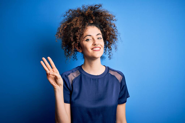 Jovem mulher bonita com cabelo encaracolado e piercing vestindo casual camiseta azul mostrando e apontando para cima com os dedos número três enquanto sorri confiante e feliz
. - Foto, Imagem