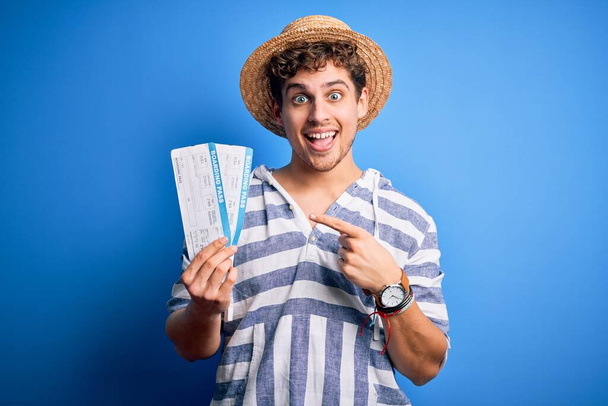 Junger blonder Touristenmann mit lockigem Haar im Urlaub mit Sommermütze und Bordkarte sehr glücklich mit Hand und Finger zeigend - Foto, Bild