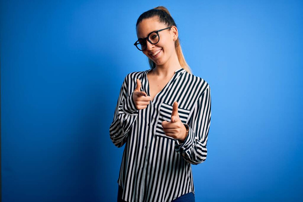 Beautiful blonde woman with blue eyes wearing striped shirt and glasses over blue background pointing fingers to camera with happy and funny face. Good energy and vibes. - Photo, Image