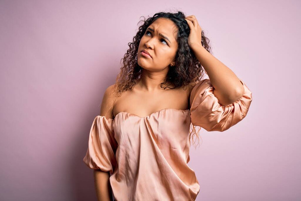 Young beautiful woman with curly hair wearing casual t-shirt standing over pink background confuse and wondering about question. Uncertain with doubt, thinking with hand on head. Pensive concept. - Foto, Imagen
