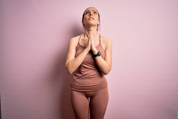 Junge blonde Fitness-Frau in Sportkleidung über rosa isoliertem Hintergrund bettelt und betet mit den Händen zusammen mit hoffnungsvollem Gesichtsausdruck sehr emotional und besorgt. Betteln. - Foto, Bild