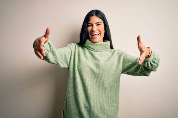 Νεαρή όμορφη ισπανόφωνη γυναίκα που φοράει πράσινο χειμωνιάτικο πουλόβερ σε απομονωμένο φόντο κοιτάζοντας την κάμερα χαμογελώντας με ανοιχτές αγκάλες για αγκαλιά. Χαρούμενη έκφραση που αγκαλιάζει την ευτυχία. - Φωτογραφία, εικόνα