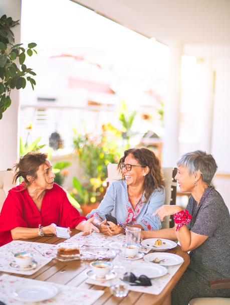 Setkání žen středního věku na obědě a popíjení kávy. Starší přátelé usměvaví šťastný pomocí smartphone doma na slunný den - Fotografie, Obrázek