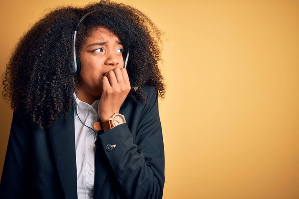 Afro saçlı, sarı arka planda kulaklık takan genç Afrikalı Amerikalı bir kadın. Ağzı tırnak yiyen elleri yüzünden gergin ve stresli görünüyor. Endişe sorunu. - Fotoğraf, Görsel