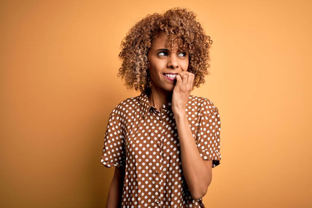 Young beautiful african american woman wearing casual shirt standing over yellow background looking stressed and nervous with hands on mouth biting nails. Anxiety problem. - Fotoğraf, Görsel
