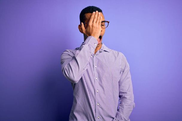 Bel homme afro-américain portant une chemise rayée et des lunettes sur fond violet bâillement fatigué couvrant la moitié du visage, des yeux et de la bouche avec la main. Le visage souffre dans la douleur
. - Photo, image