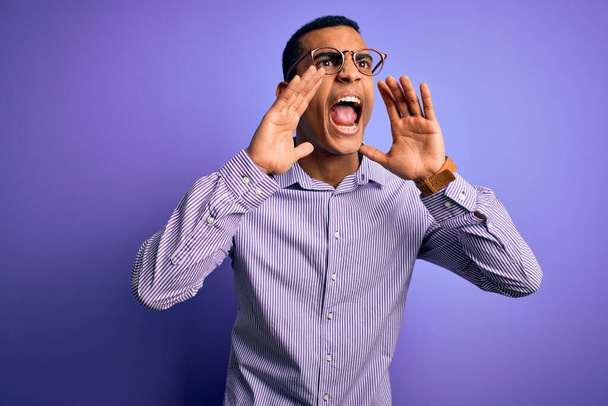 Komea afrikkalainen amerikkalainen mies yllään raidallinen paita ja lasit violetti tausta huutaa vihainen ääneen kädet suun päällä
 - Valokuva, kuva