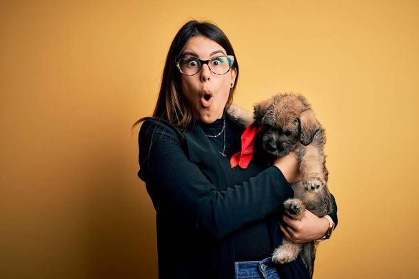 Young beautiful brunette woman holding cute puppy pet over isolated yellow background scared in shock with a surprise face, afraid and excited with fear expression - Photo, Image
