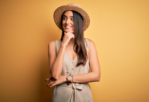 Young beautiful brunette woman on vacation wearing casual dress and hat with hand on chin thinking about question, pensive expression. Smiling with thoughtful face. Doubt concept. - Photo, Image