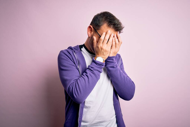 泣きながら手で顔を覆う悲しい表情でピンクの背景に立つ紫のスウェットシャツと眼鏡を身に着けている若いハンサムな男。うつ病の概念. - 写真・画像