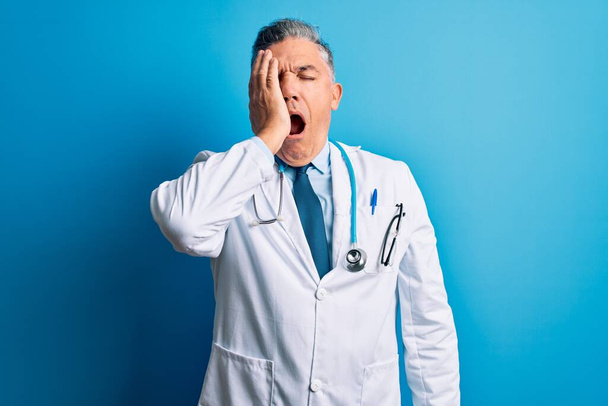 Keski-ikä komea harmaa tukka lääkäri mies yllään takki ja sininen stetoskooppi Haukottelu väsynyt kattaa puoli kasvot, silmä ja suu käsin. Kasvot kipeä kipu
. - Valokuva, kuva