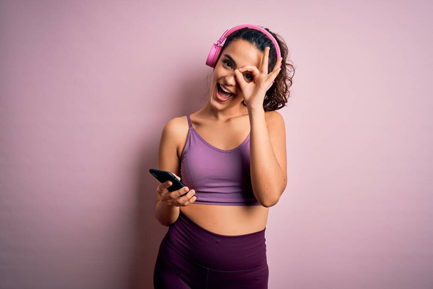 Sportvrouw met krullend haar doen sport luisteren naar muziek met behulp van smartphone en hoofdtelefoon met gelukkig gezicht glimlachen doen ok teken met de hand op oog kijken door vingers - Foto, afbeelding