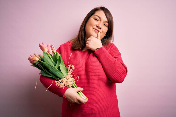 Schöne Plus-Size-Frau, die einen romantischen Strauß natürlicher Tulpenblüten über rosa Hintergrund hält und selbstbewusst in die Kamera lächelt, mit verschränkten Armen und erhobener Hand am Kinn. Positiv denken. - Foto, Bild