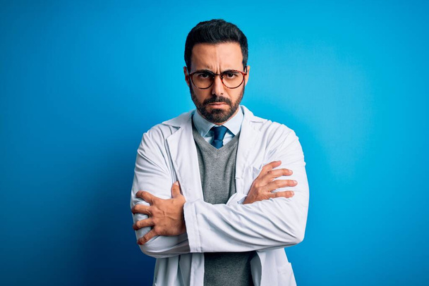 Jonge knappe dokter man met baard dragen jas en bril over blauwe achtergrond sceptisch en nerveus, afkeurende uitdrukking op gezicht met gekruiste armen. Negatief persoon. - Foto, afbeelding