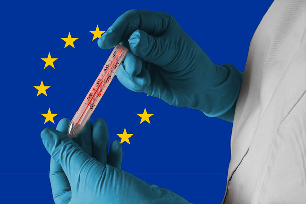Coronavirus COVID-19 járvány Európában. Az orvos az európai uniós lobogó hátterében tüdőgyulladás tüneteként magas hőmérsékletű orvosi hőmérőt tart. - Fotó, kép